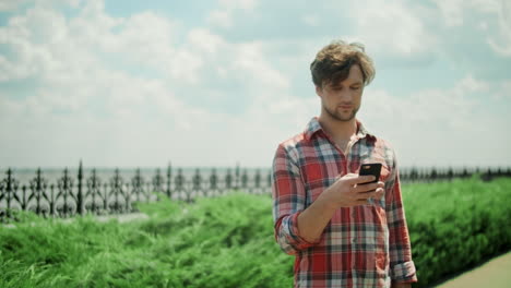 Hombre-Enfocado-Usando-Teléfono-Móvil-En-El-Parque.-Un-Tipo-Serio-Parado-En-La-Carretera-Al-Aire-Libre