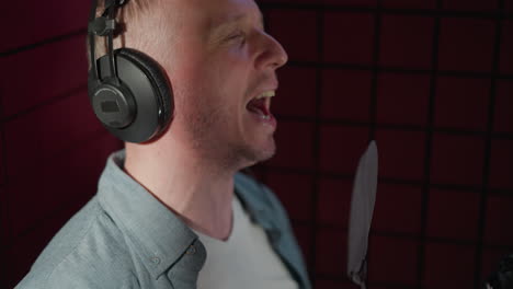 a close-up side shot of a man passionately singing with his mouth wide open and eyes closed. he is wearing black headphones and is set against a red background in a recording studio