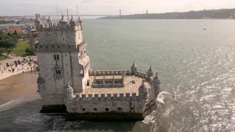 tourists traveling on belem tower near the shore of tagus river with 25 de abril bridge in background in lisbon, portugal