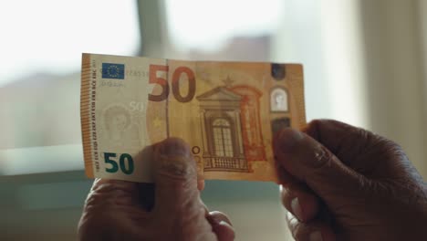 wrinkled hands of old man holding 50 euro banknote on window light background, senior man checking paper currency, suspicion of fraud. concept of dirty money, money laundering