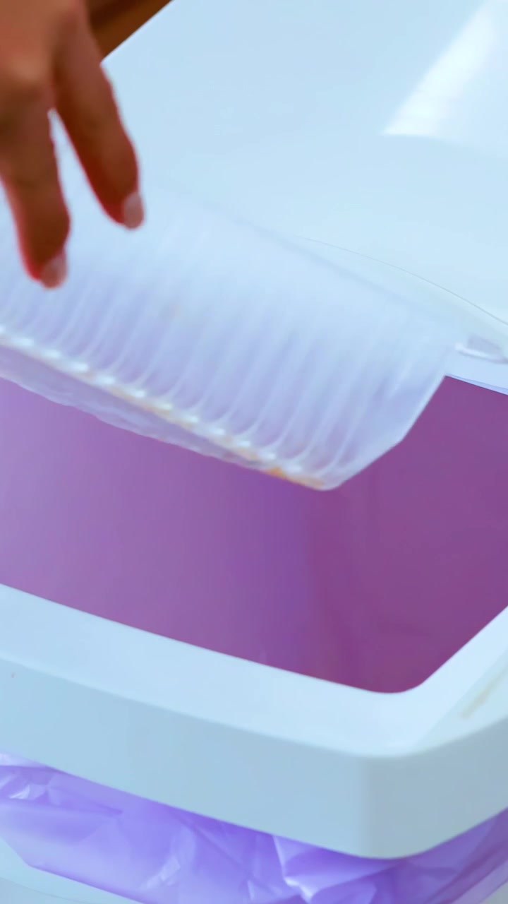 Una persona getta un contenitore vuoto di plastica per alimenti in un cestino bianco con un sacchetto viola, poi chiude il coperchio in un ambiente interno ben illuminato
