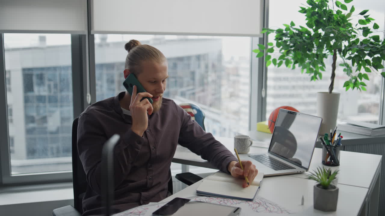 Glad freelancer ringer på mobil fra hjemmekontoret, nærbillede. Designer skriver noter.