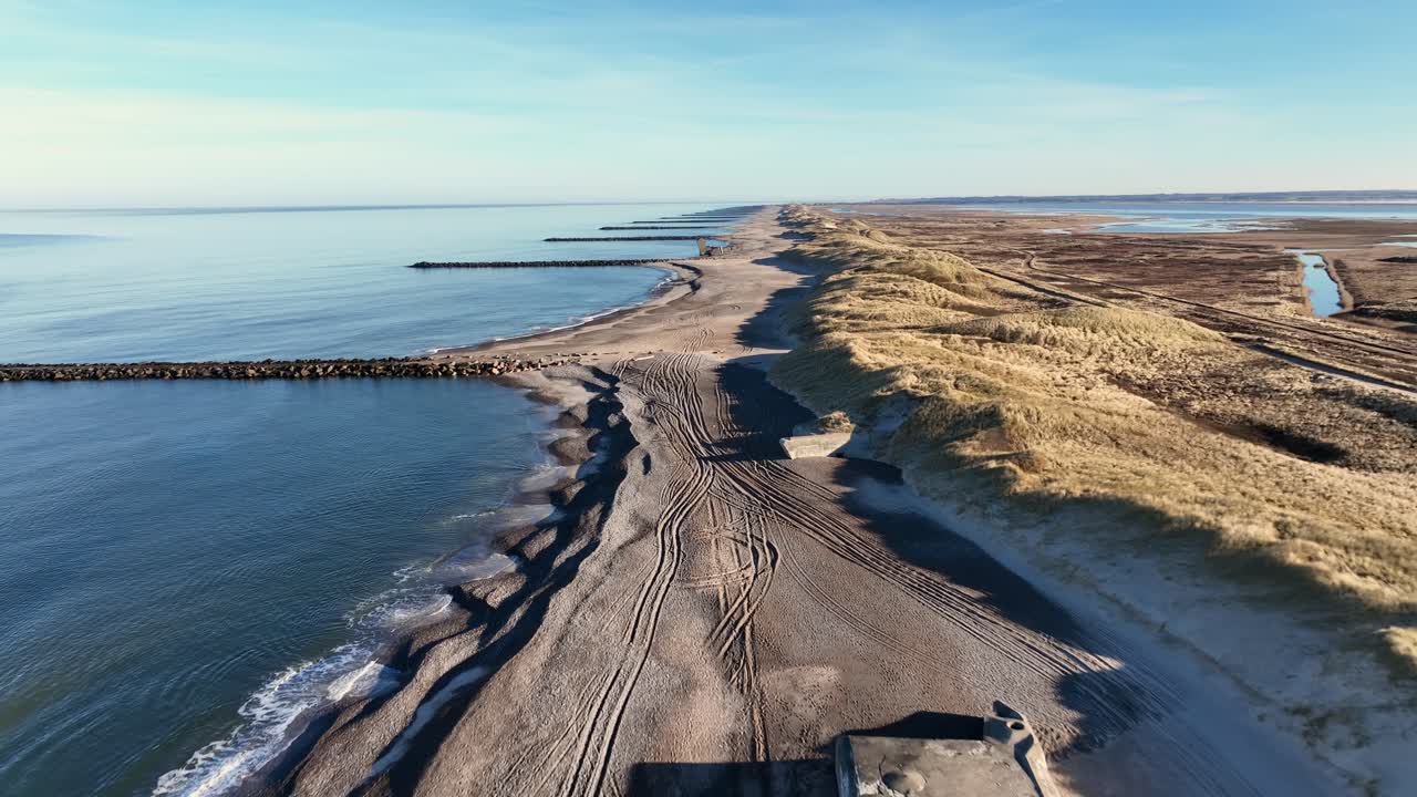 A drone captures a rugged, wild beach lined with breakwaters and dunes covered in grass. Scattered along the shoreline are weathered WWII bunkers, adding history to the remote coastal scene.