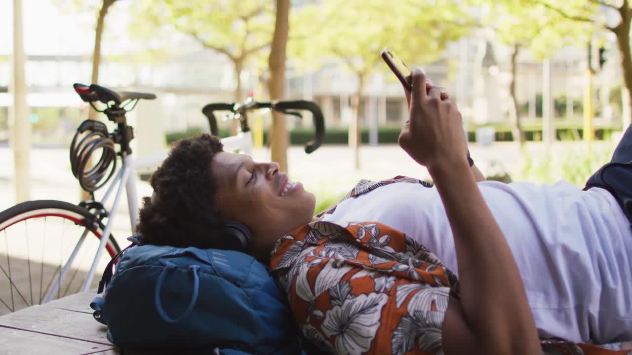 Uomo afroamericano felice in città, sdraiato su un lettino, che indossa le cuffie e usa lo smartphone al parco.