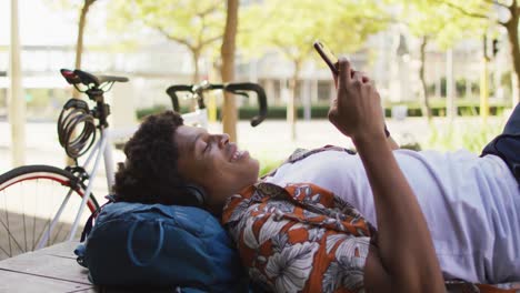 happy african american man in city, lying on deck wearing headphones and using smartphone in park