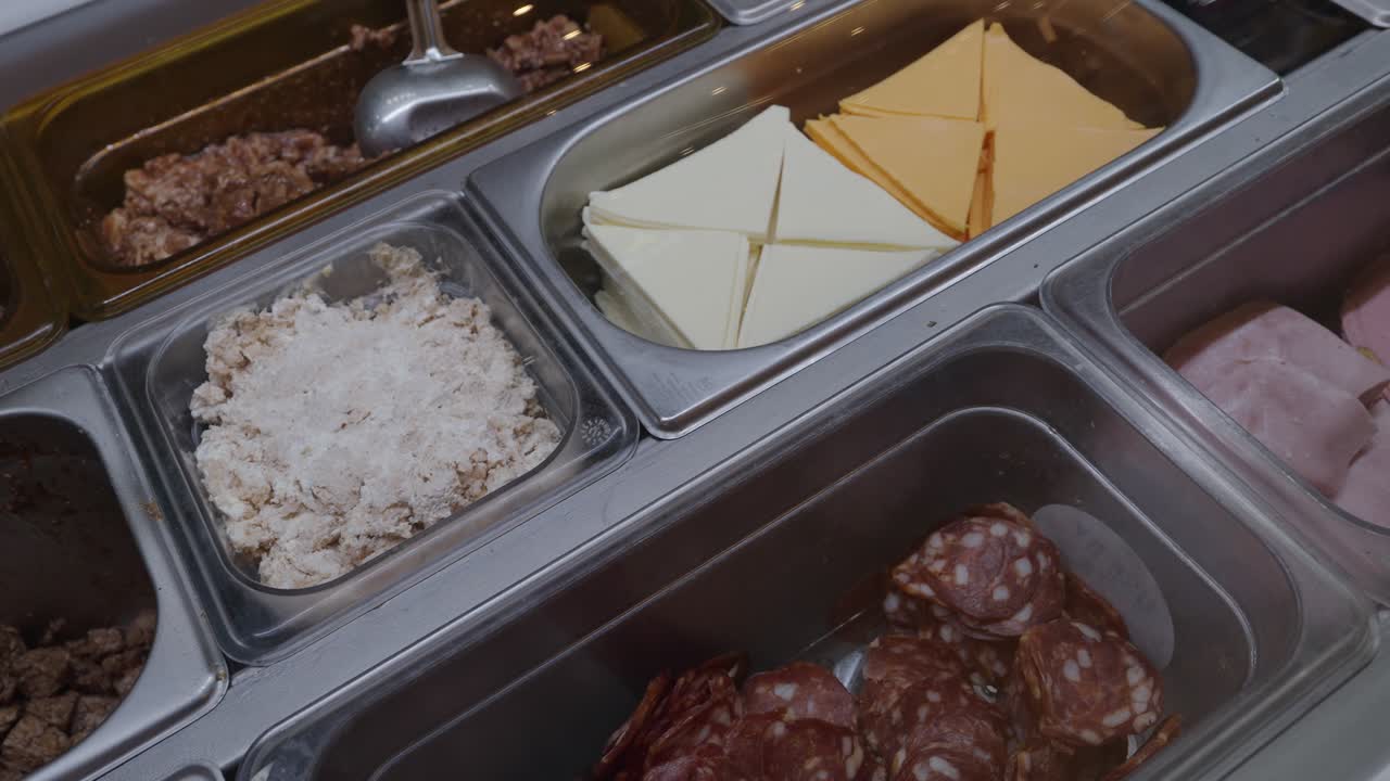Close up of stainless steel containers filled with cheese slices, ham, salami, chicken, and vegetables at a deli counter, indoor food court