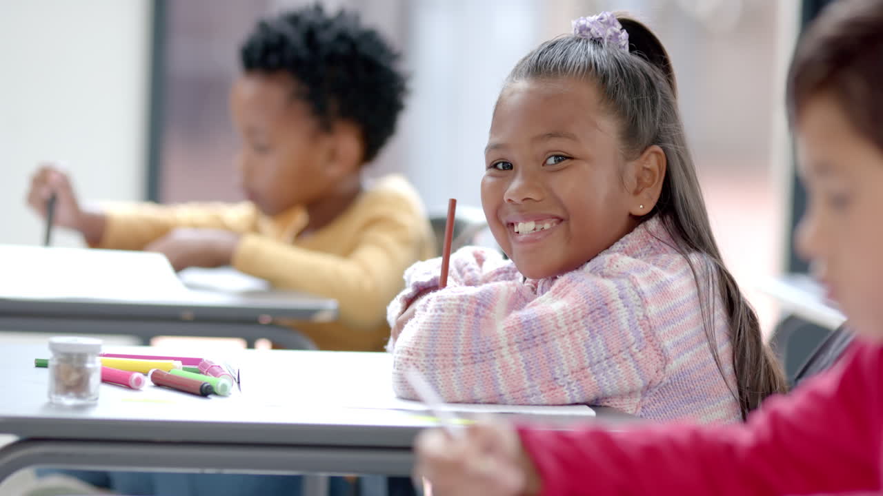 In school, smiling girl holding pencil and sitting at desk in classroom