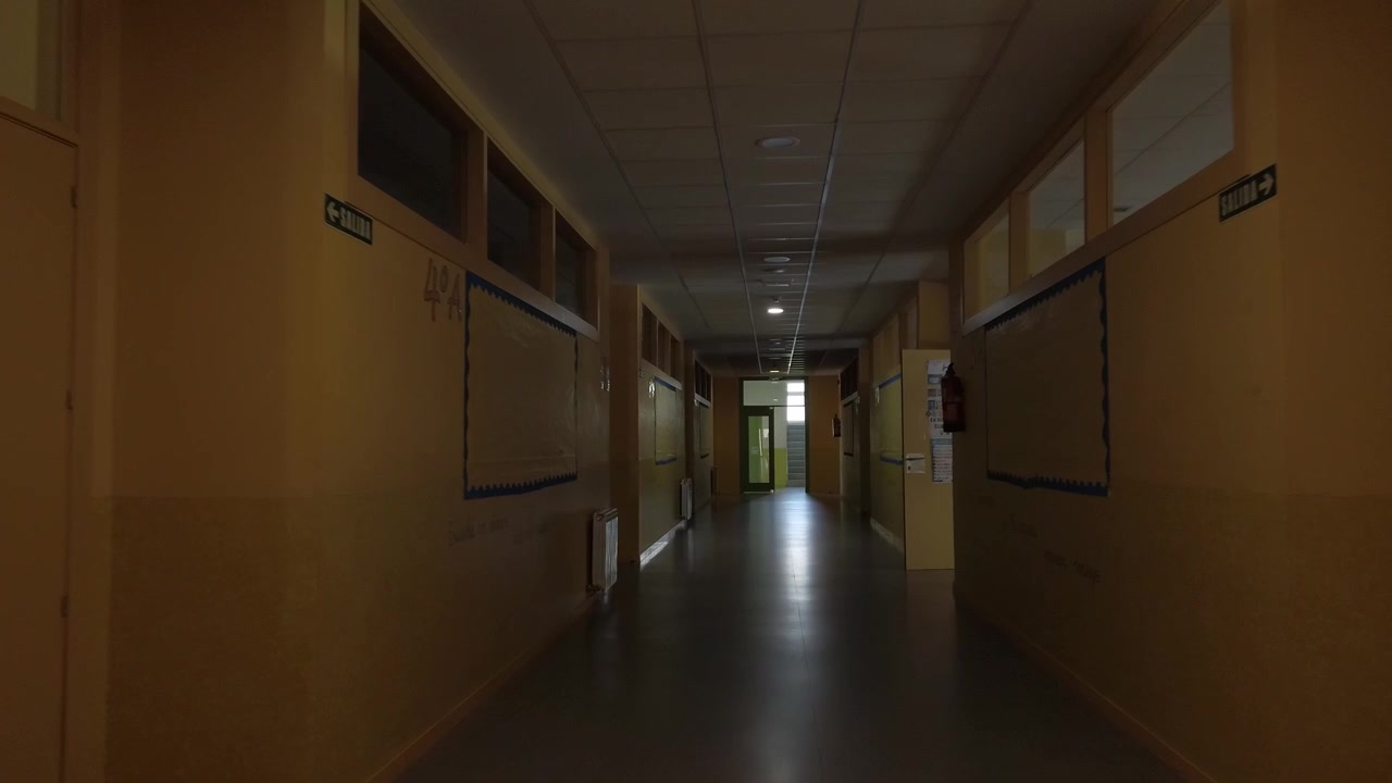 Movimento fluido della telecamera in avanti attraverso un lungo, fioco e vuoto corridoio scolastico in condizioni di scarsa illuminazione. Il corridoio buio presenta porte di aule chiuse, bacheche e una porta illuminata.