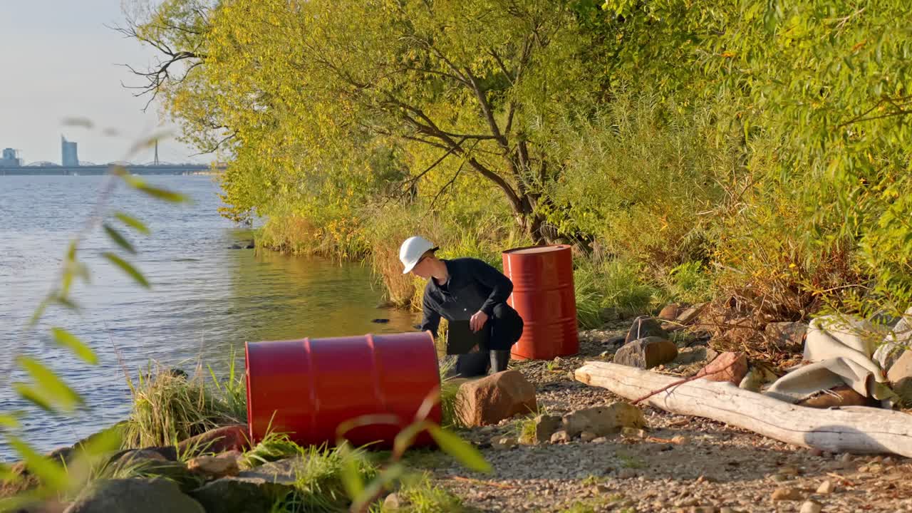 Worker in helmet testing water near red barrels along polluted riverbank in sunlight