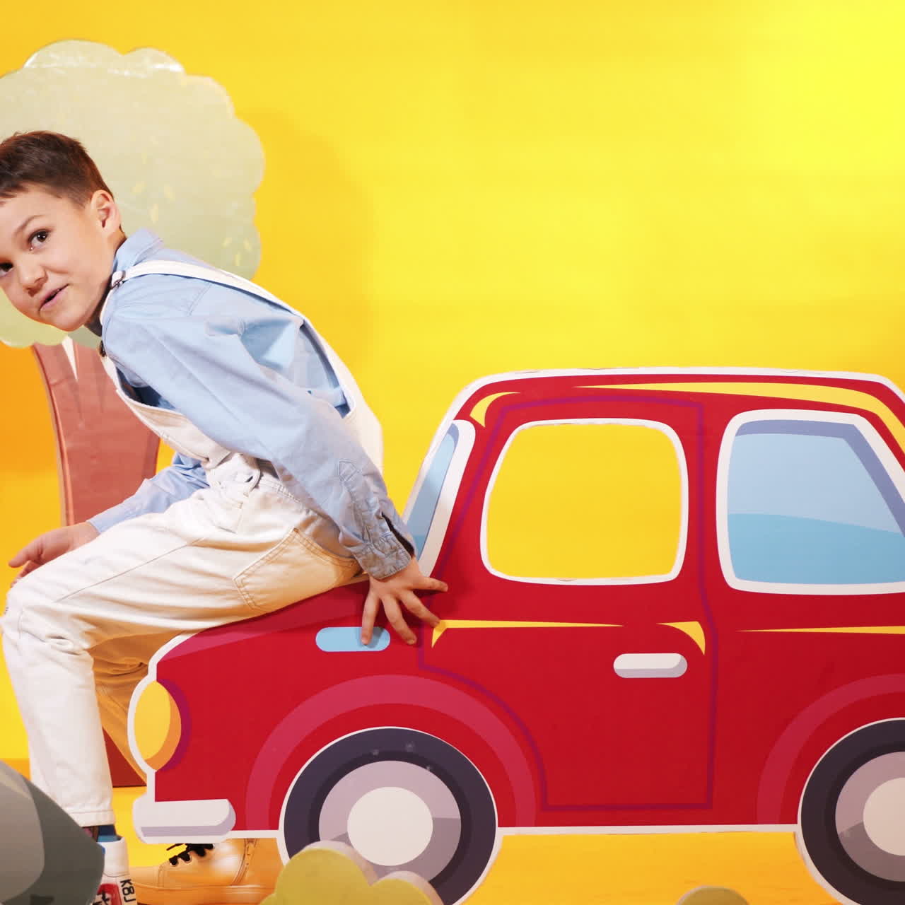 Happy kid in the children's studio with toyful decor on the yellow background. Funny little boy comes to the studio, sits on carton car and raises it up by his hands.