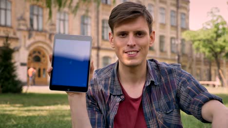 young handsome man is sitting on grass in park, taps on tablet, shows blue screen, watching at camera, relax concept, communication concept, building on background