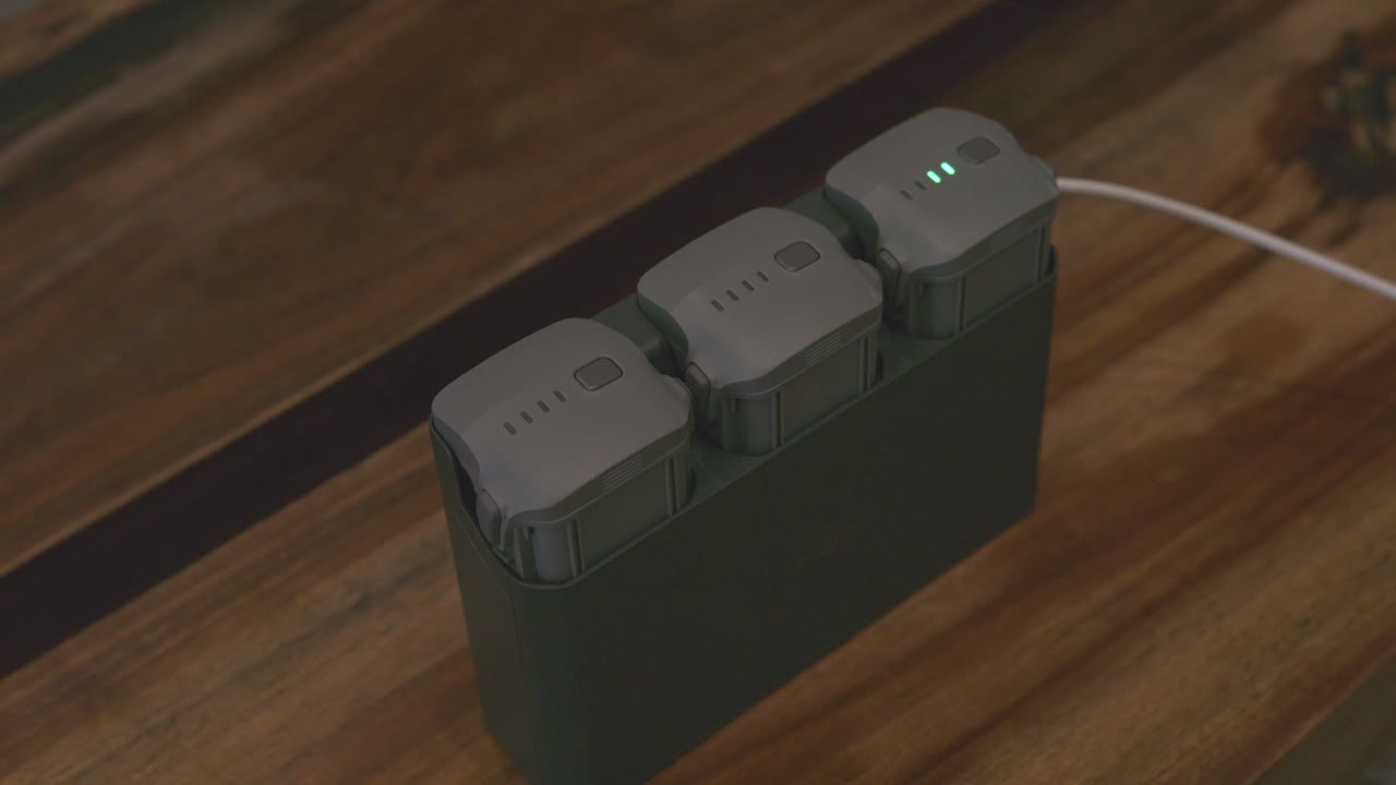 A set of three drone batteries placed in a charging dock on a wooden surface. One of the batteries shows illuminated green LED indicators, signaling an active charging process on the table