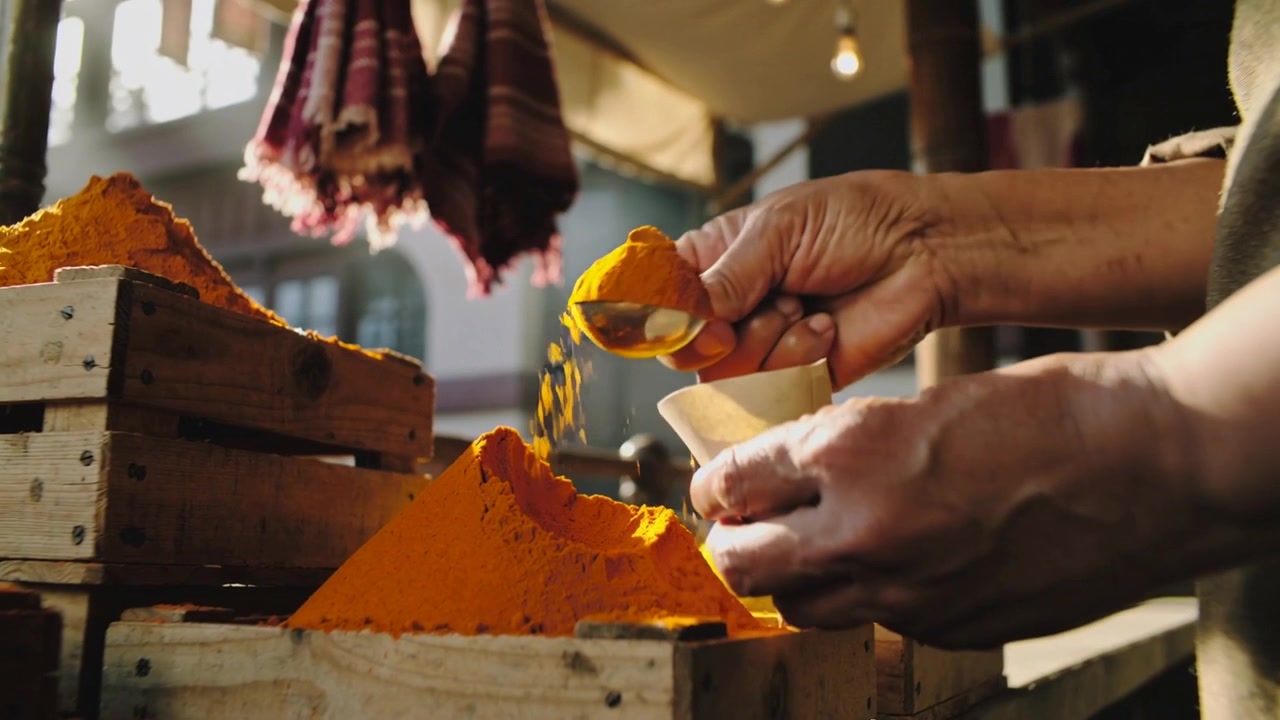 Shaping vendor hands scooping turmeric into paper cone for sale at market stall, sleeves rolled up