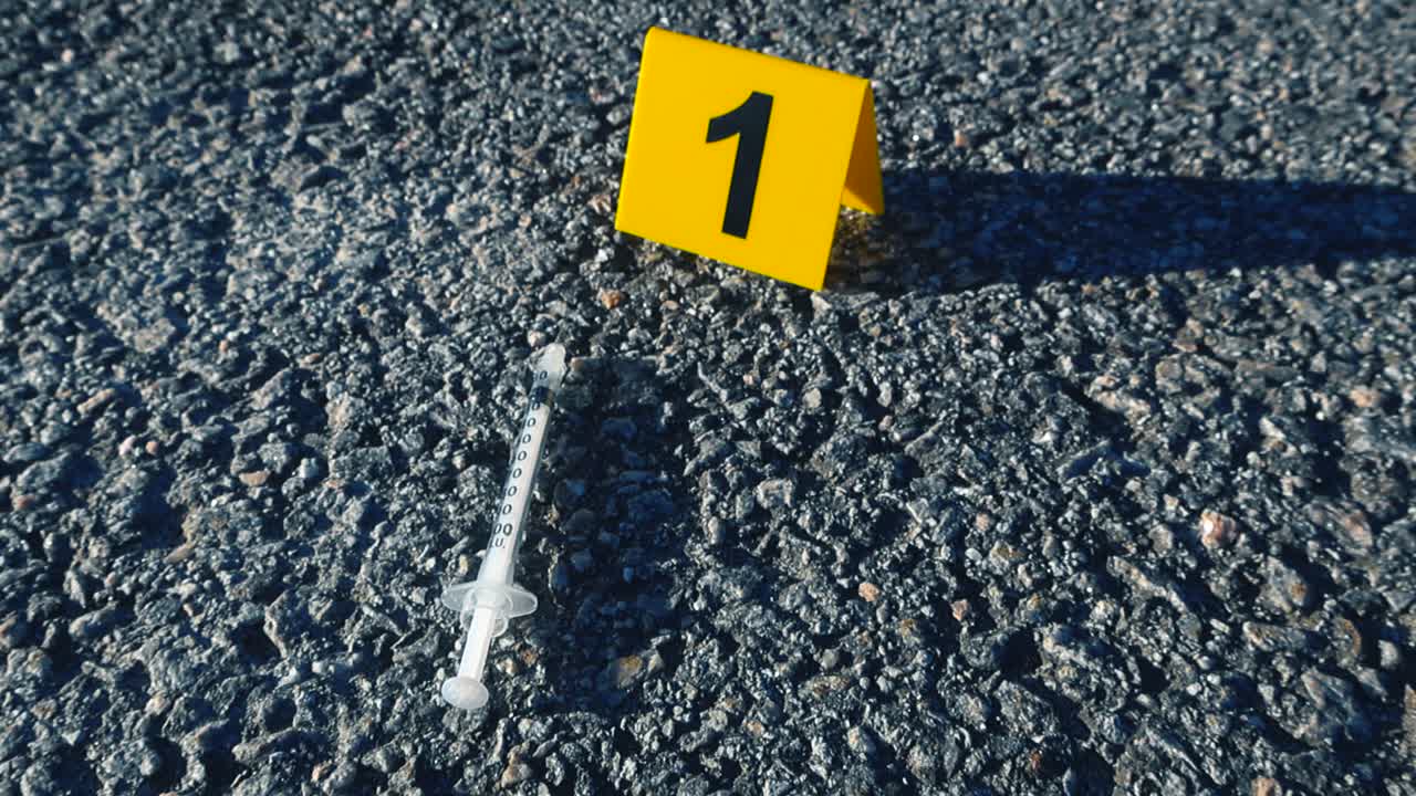 Indicatore di prova giallo con il numero uno posizionato sull'asfalto accanto a una siringa abbandonata, segnalando una scena del crimine che coinvolge l'uso di droga o una possibile overdose per indagine