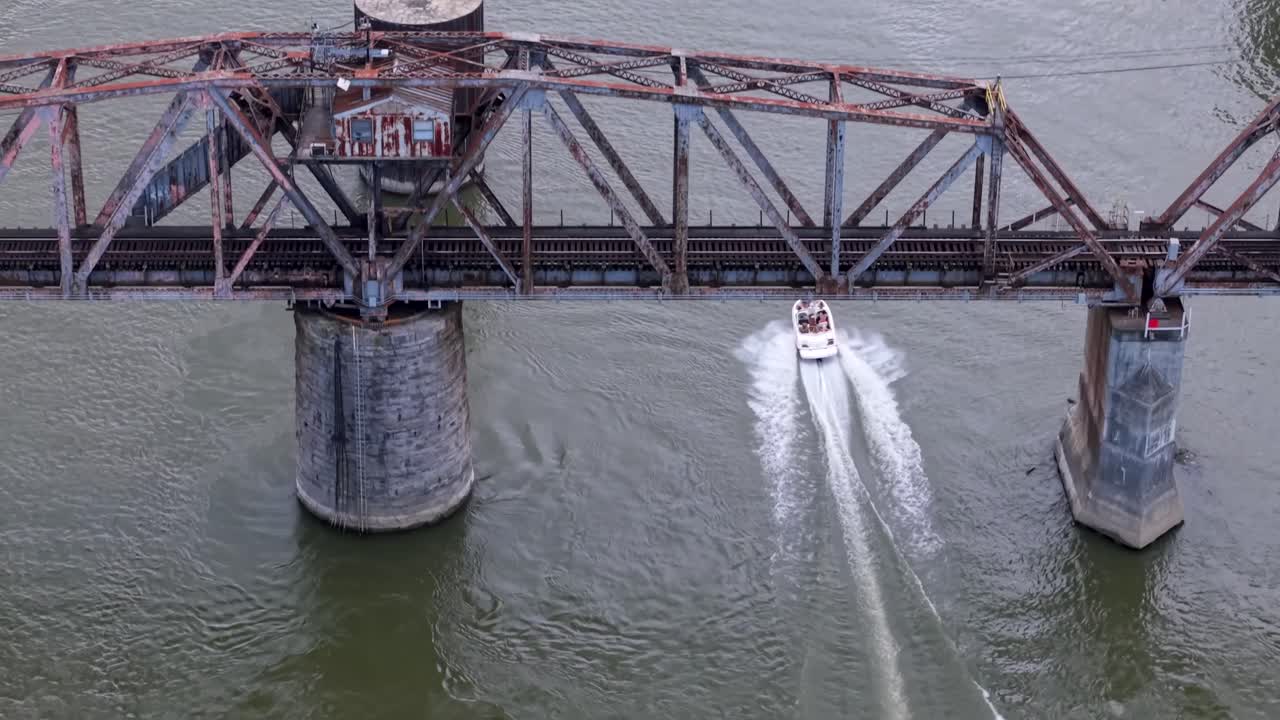Capturing boat speeding away from rustic bridge for trains as if goes down Cumberland River, aerial rising during the day