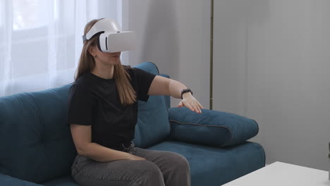 young woman is using head-mounted display for viewing interior of modern house sitting on couch in living room modern technology