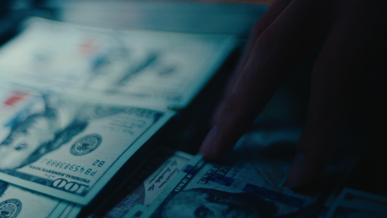 A cinematic close-up shot of a person's hand reaching for or touching a pile of one hundred dollar bills, symbolizing wealth, profit, and financial success