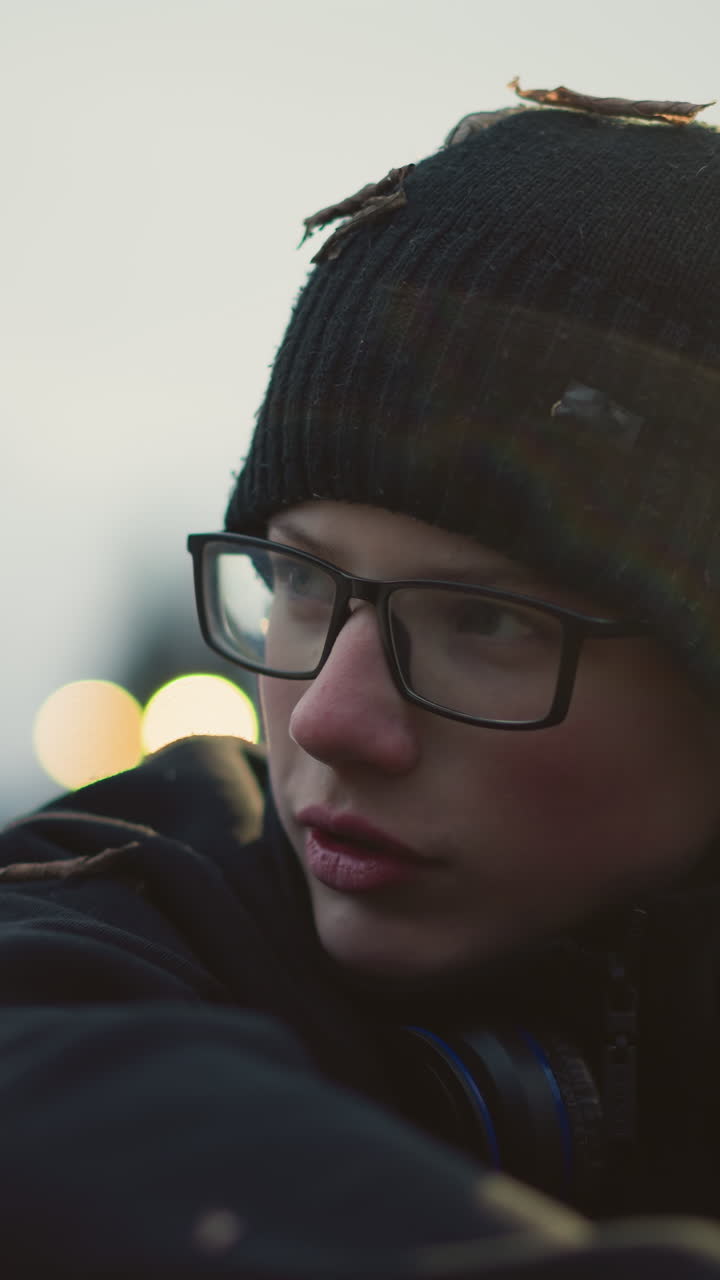 Close-up of a young boy wearing glasses and a black beanie with grass particles on his head and headphones around his neck, with light reflection on him