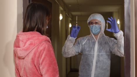 medical worker in protective suit with vaccine ampoule and syringe visiting young woman at home