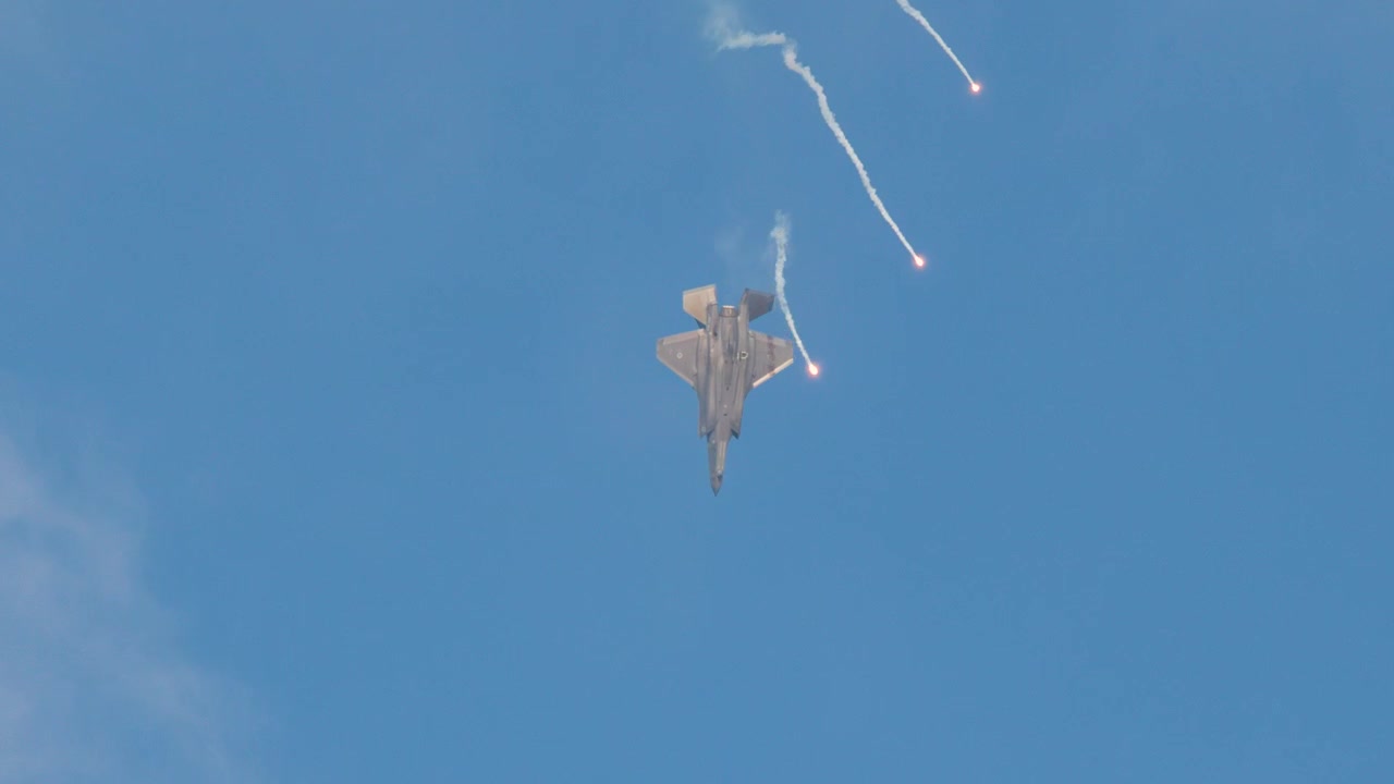 Modern stealth fighter jet performs a vertical climb and releases defensive flares in blue sky.