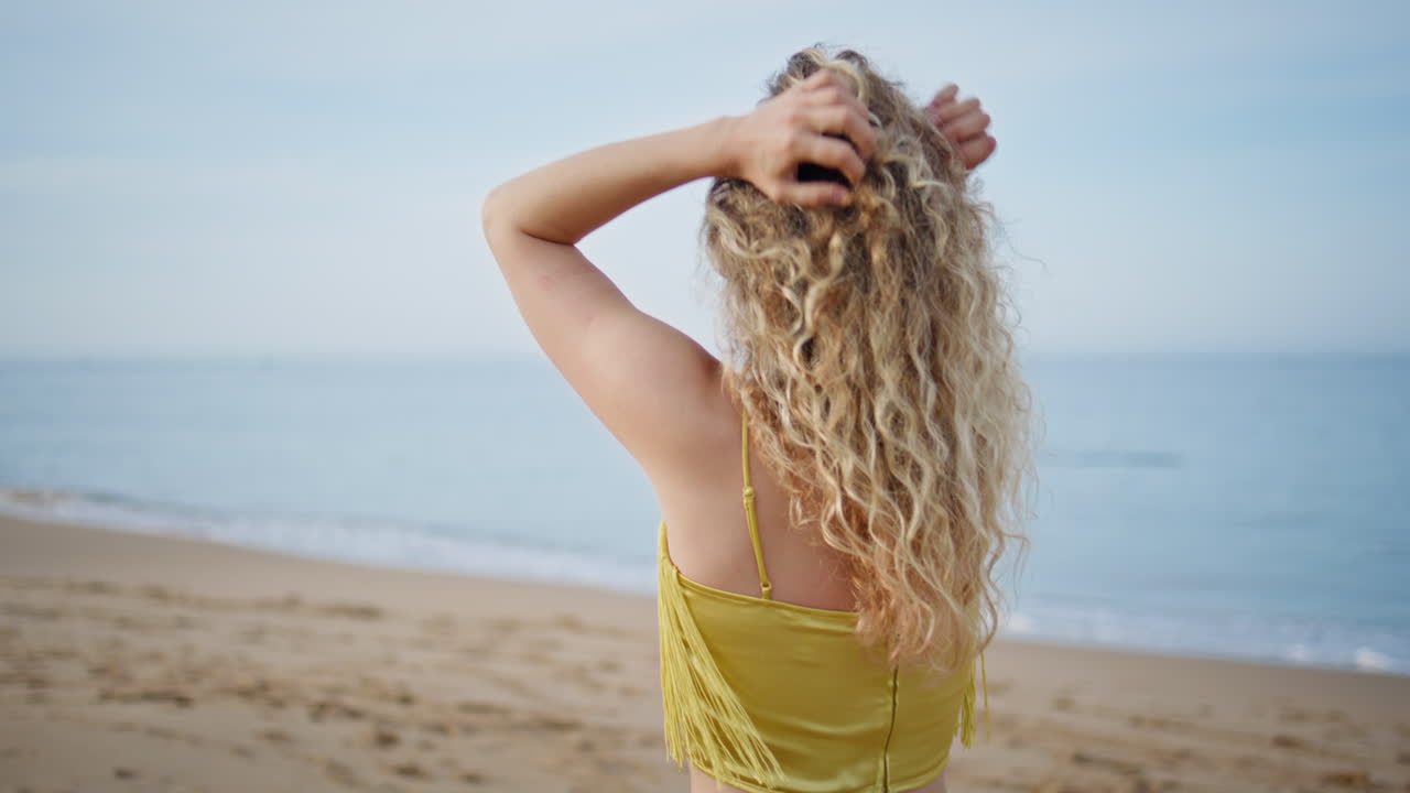 chica romántica caminando por la orilla del mar tocando el cabello rizado de cerca. vista trasera joven