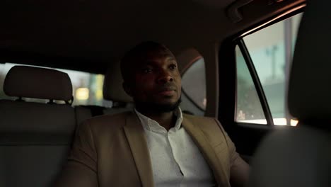 un hombre feliz y confiado con piel negra y una barba en un traje marrón viaja en un coche moderno después de una reunión de negocios y mira por la ventana