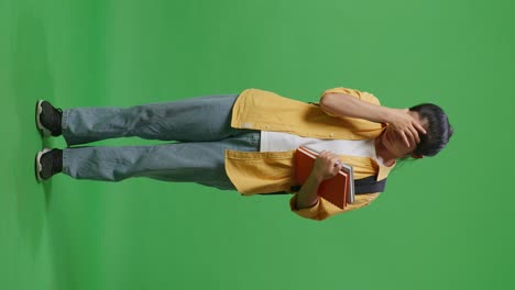 full body of asian woman student with a backpack and some books having a headache while standing in the green screen background studio