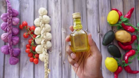 hand holding a bottle of olive oil with various vegetables in the background.