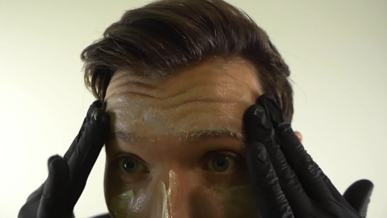 Un bell'uomo caucasico con guanti in nitrile neri applica una maschera in gel cosmetico per la cura della pelle sul viso al rallentatore, mostrando un concetto di routine di bellezza maschile su sfondo bianco