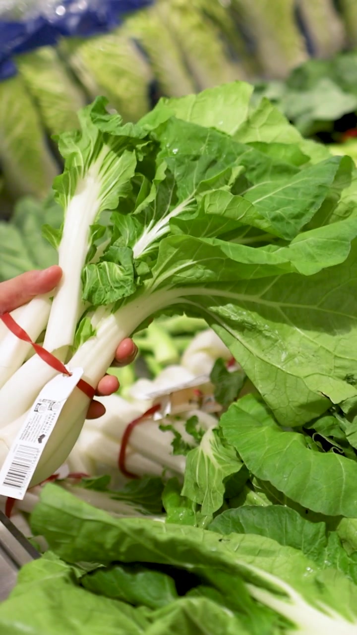 A person examines fresh bok choy in a well-lit supermarket produce section, highlighting vibrant greens and careful selection