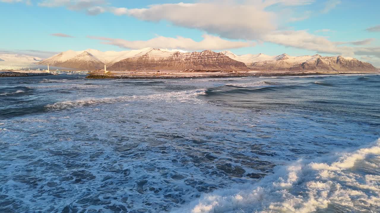 Drone view of ocean waves crashing near shoreline with distant snowy mountains, golden sunset light, scattered clouds, and lighthouse on coast creating dramatic scenic view in Iceland