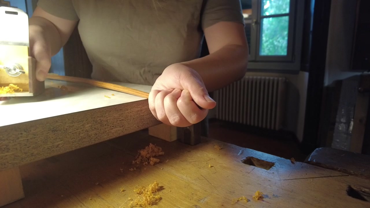 Traditional bowmaker shapes a wooden bow stave with a hand plane during the limb tapering process on a workbench, demonstrating handcrafted woodworking skills, static close up.