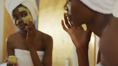 african american woman with towel watching in mirror and using cream on her face in bathroom