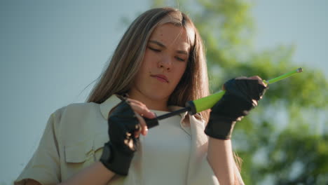 focused woman with blonde hair stands outdoors, struggling to adjust green air pump nozzle in her gloved hands, set against a natural background of lush green trees