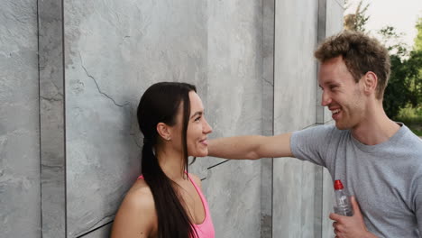 pretty runner girl leaning against a wall, drinking water and talking to her boyfriend standing in front of her while they are taking a break during training session in the city 1