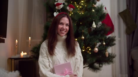 retrato de una feliz chica morena en un suéter blanco que sostiene una caja de regalos rosa cerca de un árbol de navidad decorado en una acogedora casa de invierno