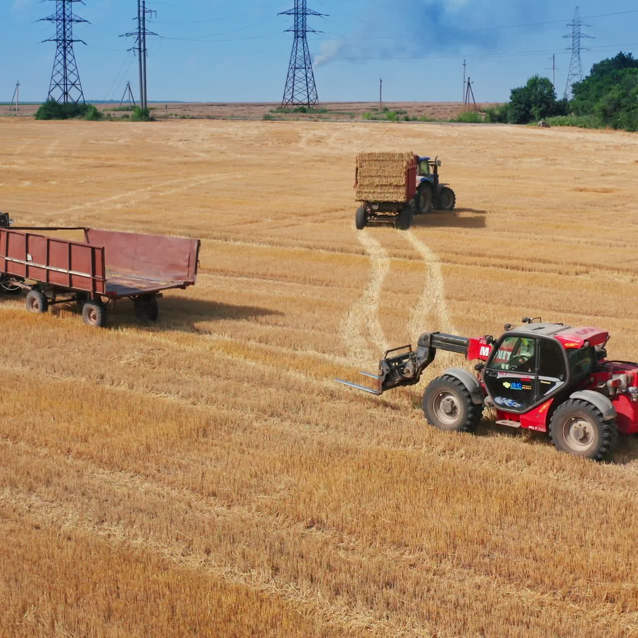Skid loader follows the empty tractor machine in the field. Loader goes to a hay bale to pick it up. Blue sky backdrop