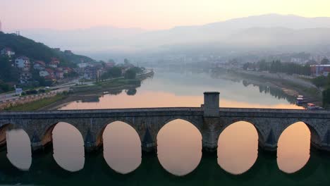 Antena-Del-Amanecer-De-Una-Persona-Caminando-Sobre-El-Puente-Del-Río-Drina-En-Visegrad,-Bosnia,