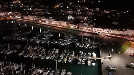 Vuelo-Nocturno-Con-Drones-Sobre-El-Puerto-Deportivo-Qe-Ii,-El-Puerto-De-San-Pedro,-Guernsey-Con-Grada,-La-Explanada-Glategny-Y-Los-Barcos-En-Sus-Amarres-Y-Las-Luces-Parpadeantes-De-La-Ciudad-Como-Telón-De-Fondo