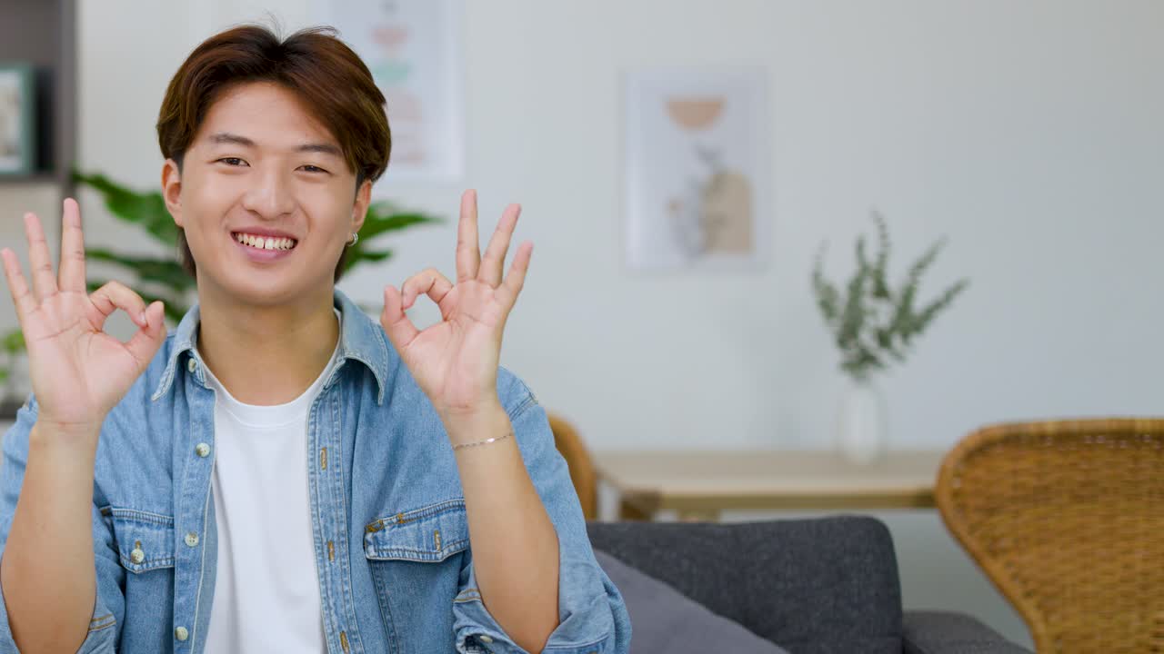 Un giovane uomo sorridente con una camicia di jeans è seduto su un divano e fa un gesto di approvazione
