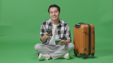 full body of asian male traveler with luggage and passport using smartphone then smiling to camera while sitting in the green screen background studio