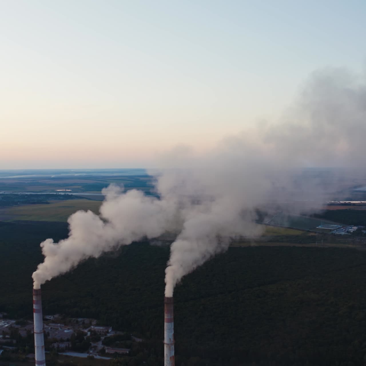 White smoke from pipes on nature background. Two chimneys produce emissions into the air among green fields and lakes.