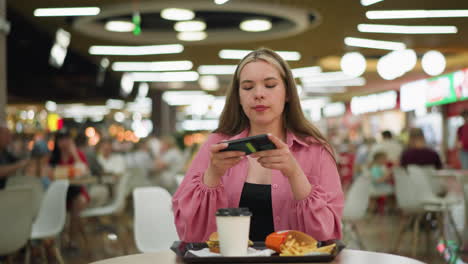 dama de vestido rosa se sienta en una mesa de madera en un restaurante bullicioso, tomando una foto de su comida, la comida incluye una hamburguesa, papas fritas y taza de café en una bandeja negra delante de ella, con un fondo borroso