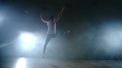 En-Un-Ballet-Moderno,-Un-Hombre-Realiza-Saltos-Y-Giros-A-La-Luz-De-Focos-Y-Humo-Sobre-Un-Fondo-Oscuro.-Ensayo-De-Coreografía-Acrobática-Del-Guión-Del-Ballet-Moderno.