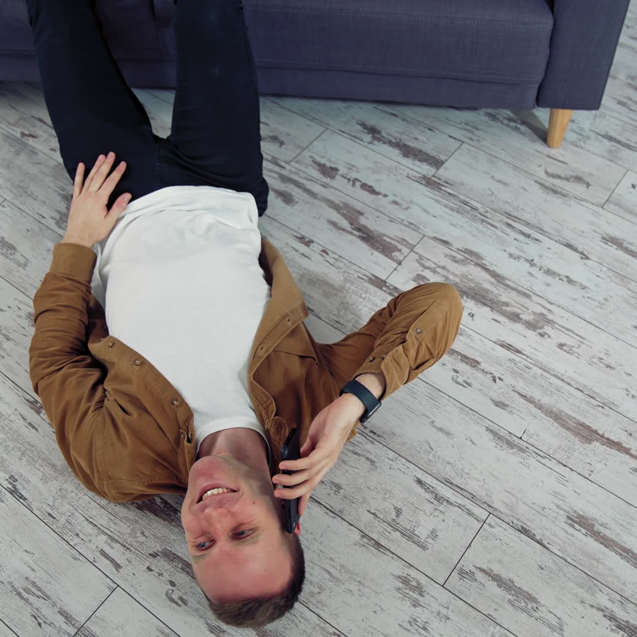 Young man talking the phone at home. Happy man having a conversation through the phone while lying on floor near the sofa. Top view. Quarantine