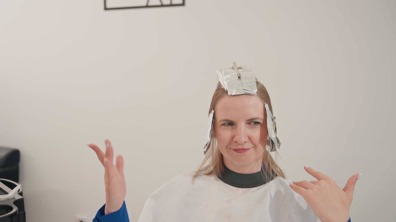 Mujer con reflejos hechos con papel de aluminio sonriendo de forma divertida. La clienta está sentada con una capa blanca durante el tratamiento de reflejos, con papel de aluminio en el pelo, gesticulando de forma expresiva con las manos. Interior de salón minimalista y acogedor con iluminación natural y estilo desenfadado.