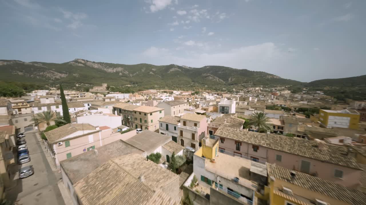 FPV Aerial drone shot over houses and church in an old town of Andratx surrounded by mountain range in Mallorca, Spain on a sunny day