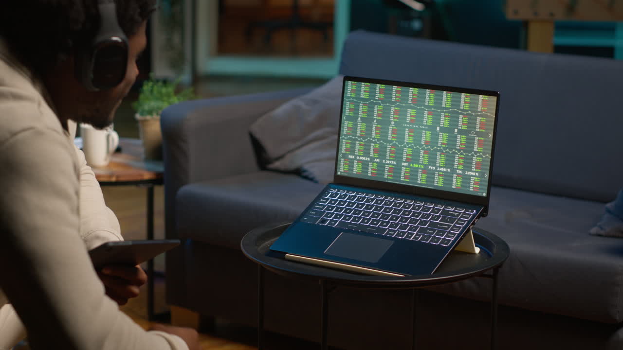Black guy listens to music and reads from tablet on the couch, waiting for stock market trends to grow and increase his income. Young adult follows financial figures on laptop, investment. Camera A.