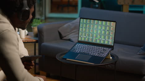 black guy listens to music and reads from tablet on the couch, waiting for stock market trends to grow and increase his income. young adult follows financial figures on laptop, investment. camera a.