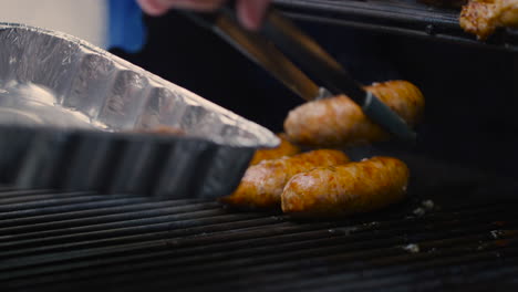 pinzas recogiendo salchichas barbacoa cocidas de la parrilla y colocándolas en una bandeja de aluminio para servirlas